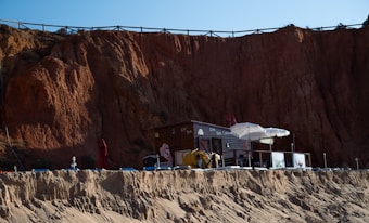 A small beachside caf&eacute; with a dark wooden exterior and white umbrellas is nestled against a towering cliff of reddish-brown rock. Sun loungers and a red parasol are visible on the sandy area in front, while the caf&eacute; has a small elevated terrace with railing.