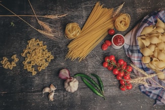 An arrangement of various types of uncooked pasta displayed alongside fresh vegetables and herbs on a rustic wooden surface. Bow tie pasta, spaghetti, and shell pasta are spread out and accompanied by cherry tomatoes on the vine, green chili peppers, garlic cloves, a halved red onion, and wheat stalks. A small dish of red tomato sauce is placed near the center, enhancing the culinary theme.