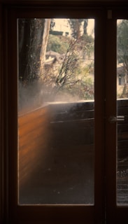 A foggy glass door with a wooden frame. Outside, there are visible blurry plants and trees, with sunlight filtering through the foliage. The wooden balcony railing is slightly visible through the fogged glass.