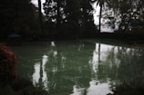 A reflective wet tennis court surrounded by tall trees and foliage, with a distant view of a cityscape partially visible through the trees. The overcast sky creates a dim and moody ambiance.