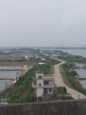 A rural landscape featuring a narrow road bordered by lush greenery, leading to a small building with multiple floors. There are bodies of water on either side of the road, possibly fish ponds or rice fields. In the background, there appears to be industrial structures, partially obscured by haze.