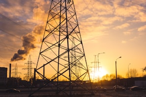 Steel electrical towers are silhouetted against a warm sunset sky, with a factory emitting smoke in the background. Cars are seen on the road and streetlights are visible, creating an industrial ambiance.