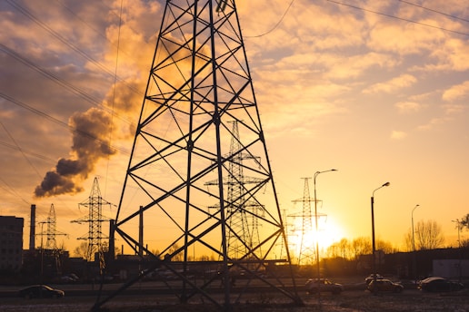 Steel electrical towers are silhouetted against a warm sunset sky, with a factory emitting smoke in the background. Cars are seen on the road and streetlights are visible, creating an industrial ambiance.