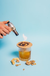 A hand holding a small blowtorch is igniting a piece of wood placed over a glass filled with an amber liquid. There are peanut shells and nuts scattered around the glass on a light blue background.