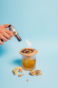 A hand holding a small blowtorch is igniting a piece of wood placed over a glass filled with an amber liquid. There are peanut shells and nuts scattered around the glass on a light blue background.