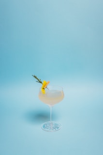 A cocktail glass filled with a pale yellow drink garnished with a sprig of rosemary and a twist of lemon peel. The glass stands on a light blue background, giving a clean and minimalist appearance.