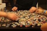 Several hands are sorting through a large number of colorful puzzle pieces scattered on a dark table. A partially open box is visible on the side.