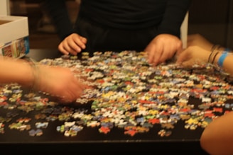 Several hands are sorting through a large number of colorful puzzle pieces scattered on a dark table. A partially open box is visible on the side.