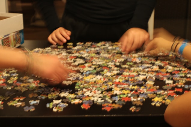 Several hands are sorting through a large number of colorful puzzle pieces scattered on a dark table. A partially open box is visible on the side.