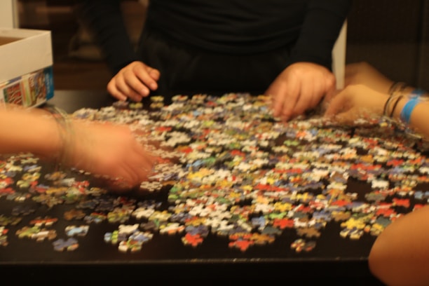 Several hands are sorting through a large number of colorful puzzle pieces scattered on a dark table. A partially open box is visible on the side.
