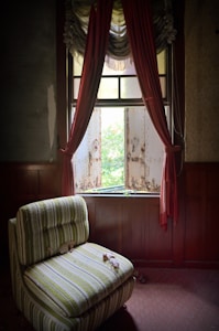 A vintage chair with green and white stripes sits in front of a window with tattered red curtains. The window is partially open, revealing rusty shutters and a glimpse of greenery outside, suggesting an abandoned or neglected space. The room has dark wood paneling and a red carpet with some leaves scattered on the floor.