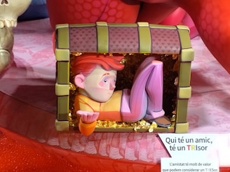 A colorful sculpture of a stylized human figure with orange hair, dressed in a yellow and pink outfit, appears to be sitting inside a large treasure chest. The chest is adorned with greenish edges and a pattern of small hearts on its exterior. The interior of the chest is filled with golden decorations. The background includes red and pink hues with part of a skull visible on the left side. A sign with text in Catalan is partially visible near the sculpture.