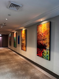 A hallway with several colorful abstract paintings hanging on the wall. The ceiling has recessed lighting, and the floor is covered with a patterned carpet. The paintings mostly feature vibrant shades of red, yellow, blue, and black, and their abstract designs create a dynamic and artistic atmosphere.