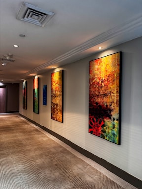 A hallway with several colorful abstract paintings hanging on the wall. The ceiling has recessed lighting, and the floor is covered with a patterned carpet. The paintings mostly feature vibrant shades of red, yellow, blue, and black, and their abstract designs create a dynamic and artistic atmosphere.
