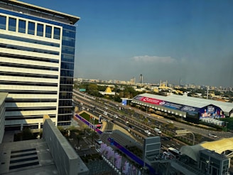A modern cityscape features a large, glass-fronted office building in the foreground. A major highway with multiple lanes and several vehicles is visible, running parallel to the building. Surrounding the highway are banners advertising a food festival. In the background, various buildings of different heights populate the skyline under a clear blue sky.