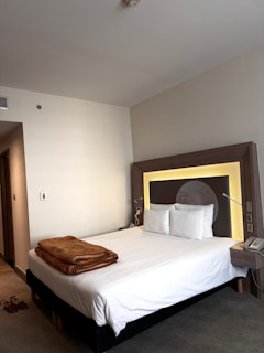 A neatly made bed with white sheets and two pillows is positioned against a headboard with illuminated backlighting. A brown blanket is folded and placed at the foot of the bed. To the right of the bed is a small table with a telephone on it. The room has neutral-colored walls and a carpeted floor. A pair of brown shoes is visible on the floor.