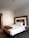 A neatly made bed with white sheets and two pillows is positioned against a headboard with illuminated backlighting. A brown blanket is folded and placed at the foot of the bed. To the right of the bed is a small table with a telephone on it. The room has neutral-colored walls and a carpeted floor. A pair of brown shoes is visible on the floor.