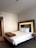 A neatly made bed with white sheets and two pillows is positioned against a headboard with illuminated backlighting. A brown blanket is folded and placed at the foot of the bed. To the right of the bed is a small table with a telephone on it. The room has neutral-colored walls and a carpeted floor. A pair of brown shoes is visible on the floor.