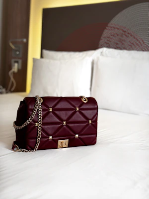 A stylish maroon quilted handbag with gold chain straps and gold stud accents is placed on a neatly made white bed. The background features a headboard with modern design elements and soft lighting that creates a cozy ambiance.