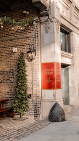 A quaint urban setting with a brick wall and an ornate stone facade. There is a festive Christmas tree adorned with lights next to a vintage-style lamp post. A red sign with the name 'Trivoli Tavern' is prominently displayed on the building's corner. Cobblestone pavement and string lights add a rustic charm to the scene.