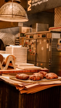 A rustic kitchen setting features a stack of plates on a counter, alongside several large cooked meats resting on a tray. Industrial elements like metal lights and equipment are visible in the background, suggesting a professional kitchen environment.
