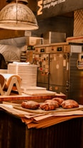 A rustic kitchen setting features a stack of plates on a counter, alongside several large cooked meats resting on a tray. Industrial elements like metal lights and equipment are visible in the background, suggesting a professional kitchen environment.