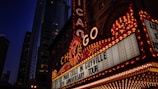 A vibrant marquee with the word 'Chicago' prominently displayed in bright lights. The marquee advertises 'Liz Phair Exile in Guyville 30th Anniversary Tour.' Tall buildings with illuminated windows form the background, suggesting an urban cityscape at night.