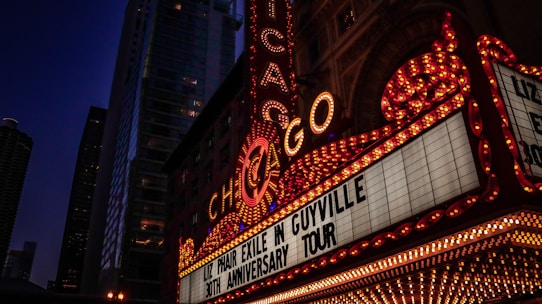 A vibrant marquee with the word 'Chicago' prominently displayed in bright lights. The marquee advertises 'Liz Phair Exile in Guyville 30th Anniversary Tour.' Tall buildings with illuminated windows form the background, suggesting an urban cityscape at night.