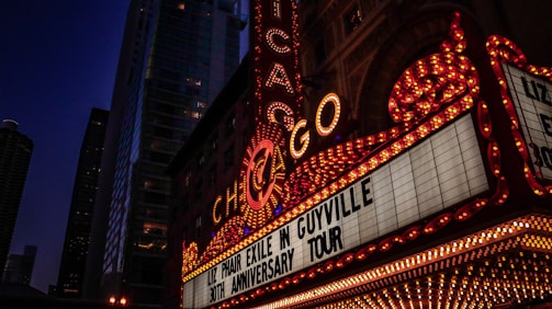 A vibrant marquee with the word 'Chicago' prominently displayed in bright lights. The marquee advertises 'Liz Phair Exile in Guyville 30th Anniversary Tour.' Tall buildings with illuminated windows form the background, suggesting an urban cityscape at night.