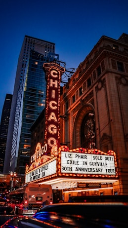 A brightly lit theater marquee is prominently displayed against an urban backdrop at dusk. The marquee features the name 'CHICAGO' in large, illuminated letters and announces a concert by Liz Phair with the text 'SOLD OUT! EXILE IN GUYVILLE 30TH ANNIVERSARY TOUR.' The ornate architectural details of the theater and adjacent towering buildings are visible under the deep blue evening sky.