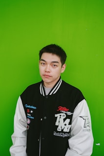 A person stands against a bright green background, wearing a black and white varsity jacket with various patches and designs. The person has short dark hair and is looking directly at the camera with a neutral expression.