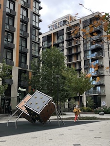 A modern urban setting features tall apartment buildings with numerous balconies. In the foreground, there is a playground with a climbing structure resembling large geometric dice. A child in a yellow jacket and red pants is running nearby. The scene is adorned with trees, some showing autumn foliage.