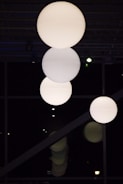 Three large, spherical light fixtures hang from the ceiling in a dark room, casting a soft glow. The background features a grid-like pattern of beams or rafters, and subtle reflections can be seen below them.
