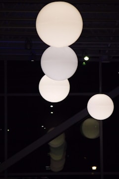 Three large, spherical light fixtures hang from the ceiling in a dark room, casting a soft glow. The background features a grid-like pattern of beams or rafters, and subtle reflections can be seen below them.