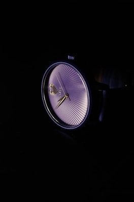 A close-up of a wristwatch with a detailed face that has radial lines emanating from the center. The watch hands are elegantly designed and the entire piece is set against a dark background, emphasizing its sleek design.