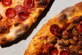 Two pizzas are displayed on a light-colored surface, each with a crispy, slightly charred crust. One pizza is topped with slices of pepperoni, while the other features sausage pieces and pepperoni.