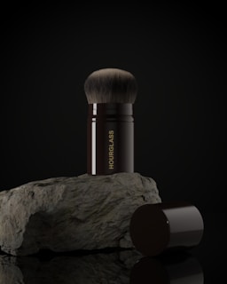 A makeup brush with a black handle and soft bristles is placed upright on a rough-textured rock. The word 'HOURGLASS' is written vertically on the handle in gold letters. The image background is dark, highlighting the brush and rock.