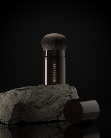 A makeup brush with a black handle and soft bristles is placed upright on a rough-textured rock. The word 'HOURGLASS' is written vertically on the handle in gold letters. The image background is dark, highlighting the brush and rock.