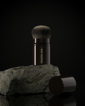 A makeup brush with a black handle and soft bristles is placed upright on a rough-textured rock. The word 'HOURGLASS' is written vertically on the handle in gold letters. The image background is dark, highlighting the brush and rock.