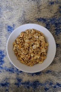 A white bowl filled with granola consisting of oats and assorted nuts, placed on a textured blue and gray patterned surface.