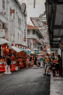 A bustling street market scene featuring numerous people walking and shopping. Stalls with vibrant red and blue canopies line the street, displaying various goods. The architecture includes traditional style buildings with ornate balconies. Hanging lights and signs with Chinese characters add cultural context, and the street is packed with a lively crowd, adding to the market atmosphere.