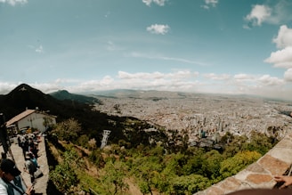 An expansive cityscape view with a dense cluster of buildings stretching towards the horizon under a blue sky scattered with clouds. The foreground features a stone terrace with people observing the scenery, surrounded by lush greenery and mountains in the background.