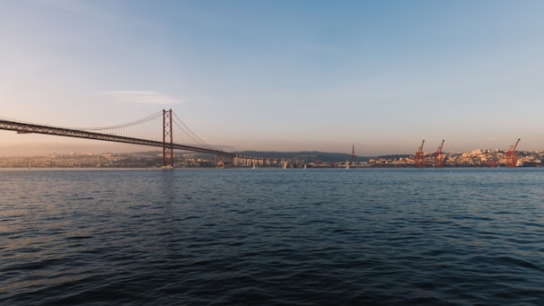 A large suspension bridge stretches across the horizon, spanning a wide body of water. The bridge connects two landmasses, with structural towers and cables visible. On the right side, several red cranes are seen at a port area, with a cityscape in the background. The sky is clear with a warm, golden hue suggesting a sunset, casting a serene light on the scene.