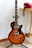 A sunburst electric guitar stands on a tripod stand with a red rotary phone resting on a wooden desk in the background.