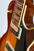 A close-up view of a portion of an electric guitar, focusing on the neck and the body. The guitar has a warm brown finish with contrasting cream and black accents, featuring metal strings and a pickup with visible screws. The image highlights the craftsmanship and details of the instrument.