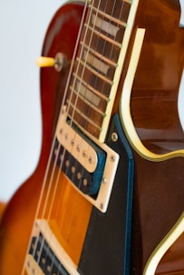 A close-up view of a portion of an electric guitar, focusing on the neck and the body. The guitar has a warm brown finish with contrasting cream and black accents, featuring metal strings and a pickup with visible screws. The image highlights the craftsmanship and details of the instrument.