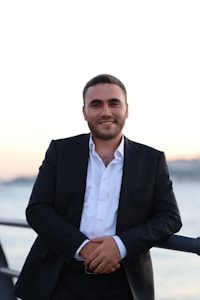 A man in a dark suit and white shirt stands by a railing with a body of water in the background. The lighting suggests a sunset or early evening.