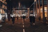 A bustling urban scene featuring a crowd of people walking through a well-lit corridor surrounded by modern architecture. Glass and metal structures are prominent, reflecting the ambient lights. There's a central train station at the end of the walkway, indicating public transport connectivity.
