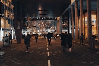 A bustling urban scene featuring a crowd of people walking through a well-lit corridor surrounded by modern architecture. Glass and metal structures are prominent, reflecting the ambient lights. There's a central train station at the end of the walkway, indicating public transport connectivity.