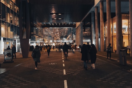 A bustling urban scene featuring a crowd of people walking through a well-lit corridor surrounded by modern architecture. Glass and metal structures are prominent, reflecting the ambient lights. There's a central train station at the end of the walkway, indicating public transport connectivity.